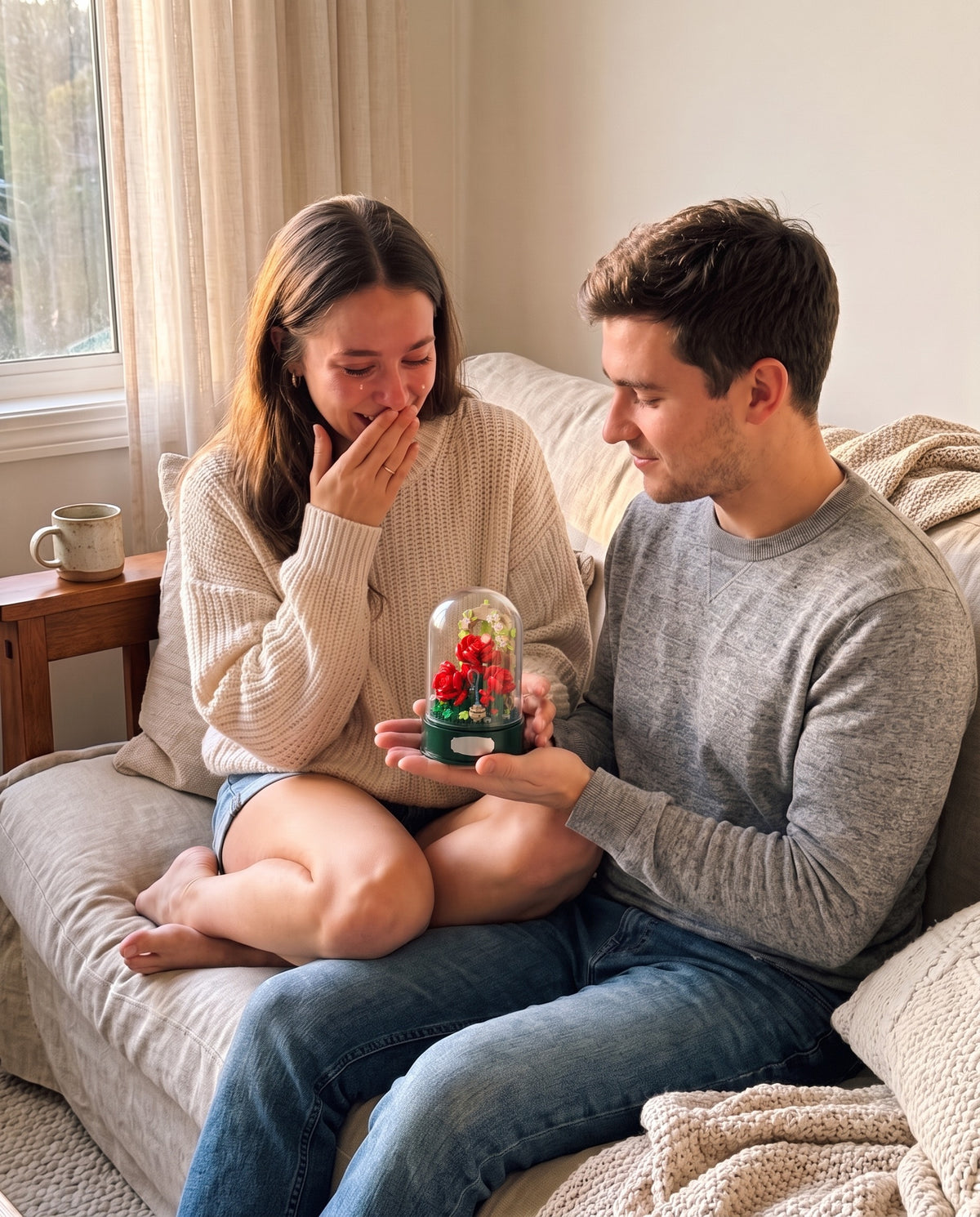 A young man handing the rose garden music box to his girlfriend on a cozy couch, her hand over her mouth