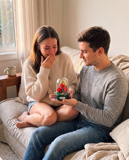 A young man handing the rose garden music box to his girlfriend on a cozy couch, her hand over her mouth