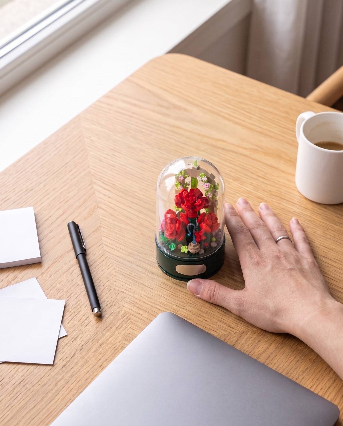 The rose garden music box on a desk next to a laptop and a mug, a hand resting beside it