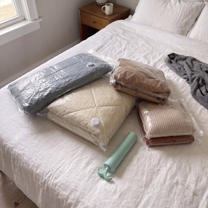 Four sealed clear vacuum storage bags of a comforter, duvet, coat and sweaters on a made bed, with the mint hand pump